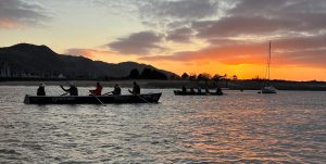 two celtic longboats rowing on the Conwy estuary, and a moored yacht, set against an orange sunset